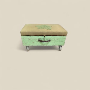 Könnte beinhalten: Kleine, rechteckige grüne Holz-Aufbewahrungsbank mit einem beige gepolsterten Oberteil. Das Oberteil hat eine grüne Blattanwendung. Die Bank hat einen dunklen Griff und steht auf vier kleinen Rollen.