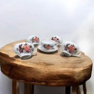 Könnte beinhalten: Ein Set aus fünf weißen Porzellan-Teetassen und Untertassen. Jede Tasse und Untertasse hat ein Blumendesign mit rosa und orangefarbenen Blüten und braunen Blättern. Die Tassen sind auf einem runden Holztisch angeordnet.