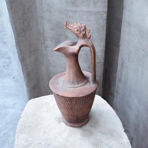 Puede incluir: Jarra de cerámica color terracota con una cabeza de dragón en el asa. La jarra tiene una base ancha con ranuras verticales y un pico. Se exhibe sobre una superficie de piedra contra un fondo neutro.