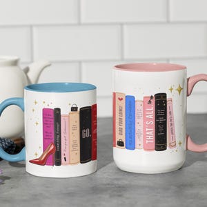 May include: Two white ceramic mugs with colorful interior and handles. One has a blue interior and handle, the other has a pink interior and handle. Each mug features a design of stacked books with text, including "Something funny" and "THAT'S ALL."