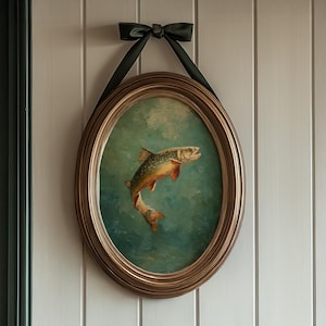 May include: An oval-shaped framed painting of a fish in shades of orange, yellow, and green against a teal background. The gold-colored frame is suspended by a black ribbon tied in a bow at the top.