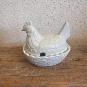 May include: A white ceramic lidded dish shaped like a hen sitting on a nest. The lid features a sculpted chicken with detailed feathers, while the base is designed to resemble a woven basket. A small notch in the rim allows for a spoon to rest.