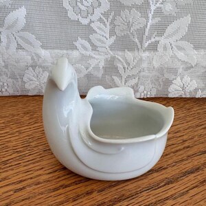 May include: A white ceramic bird-shaped dish with a smooth, glossy finish. The vessel features a stylized head and neck extending from one side, with a curved, open bowl section for holding small items. The design is minimalist and monochromatic.