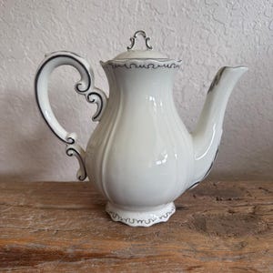 May include: A white porcelain teapot featuring a ribbed body, a curved handle, and a matching lid. The teapot is decorated with thin, dark gray or black decorative trim along the edges of the handle, the lid, the spout, and the base.