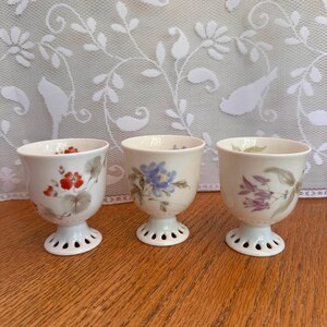 May include: Three off-white ceramic egg cups with pedestal bases featuring scalloped edges. Each cup is decorated with a different hand-painted floral motif: one with red flowers, one with blue flowers, and one with purple flowers and green leaves.