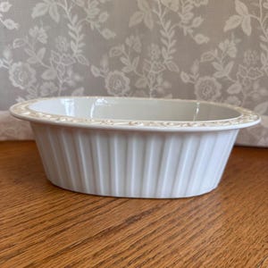 May include: An oval white ceramic baking dish featuring vertical fluted sides and a decorative embossed scroll pattern along the wide, flat rim. The interior of the dish is smooth and deep, designed for oven use with a clean, glossy finish.