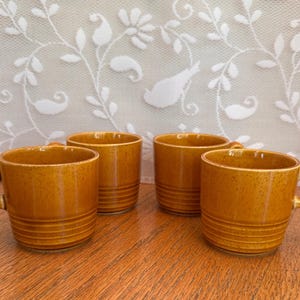 May include: Four mustard yellow ceramic mugs with a speckled finish and three horizontal ridges near the base. Each mug features a curved handle on the side. The mugs are arranged in a group, showcasing their uniform color and cylindrical shape.