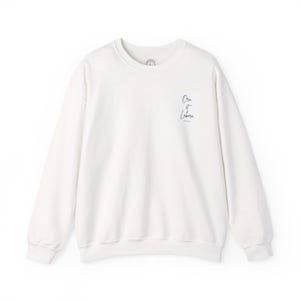 May include: A white crewneck sweatshirt with long sleeves. The text "Ora et Labora" is printed in a delicate script on the upper right chest area. The sweatshirt is made of a soft material.