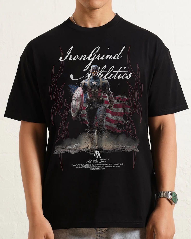 Puede incluir: Camiseta negra con un dise&ntilde;o de Capit&aacute;n Am&eacute;rica con su escudo y la bandera estadounidense. La camiseta tiene el texto "IronGrind Athletics" y "Sit The Tone", junto con un mensaje motivacional sobre el bienestar y la determinaci&oacute;n.