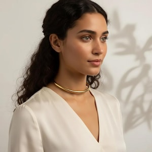 Może przedstawiać: Złoty naszyjnik typu choker. Naszyjnik ma prosty, gładki, zaokrąglony kształt tuby. Modelka ma na sobie białą bluzkę.