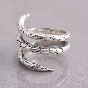 May include: A silver-colored ring in the shape of a coiled snake. The ring wraps around in a spiral design, with a textured, scale-like pattern covering the entire surface. The snake's head and tail are visible, adding detail to the jewelry.