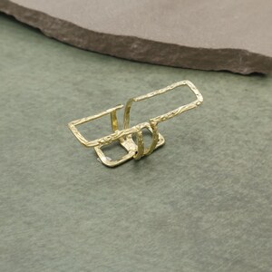 Könnte beinhalten: Ein goldfarbener Ring mit strukturierter Oberfläche. Der Ring hat ein rechteckiges Design mit zwei parallelen Stäben und einem Verbindungssteg. Der Ring befindet sich vor einem grünen Hintergrund.