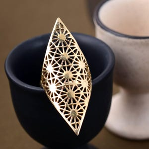 May include: A gold-colored statement ring with an openwork design. The ring features a large, elongated shape with a geometric pattern of stars and triangles. The ring is set against a dark blue background.