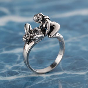 Puede incluir: Un anillo de metal plateado con un diseño escultórico de dos figuras en un abrazo íntimo. La banda es lisa y pulida, mientras que la sección superior muestra figuras detalladas y texturizadas con acentos oxidados para resaltar la forma.