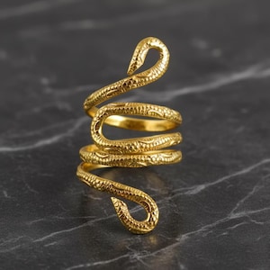 May include: A gold-colored ring in the shape of a snake, coiled around itself. The ring has detailed scale patterns and is set against a dark, marbled background. The snake's head and tail curve upwards.