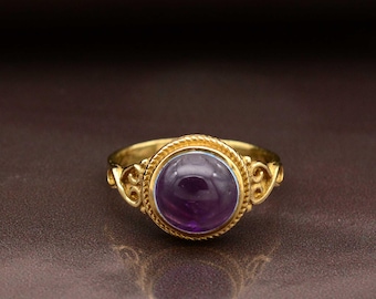 May include: A gold ring featuring a large, round, deep purple gemstone. The ring has an ornate gold band with decorative filigree on the sides and a rope-like border around the gemstone. The ring is set against a brown background.