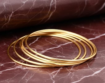 May include: A set of five thin, circular gold-toned bangles stacked together. Each bangle features a smooth, polished finish with a consistent round profile, creating a layered appearance as they overlap in a cluster on a flat surface.