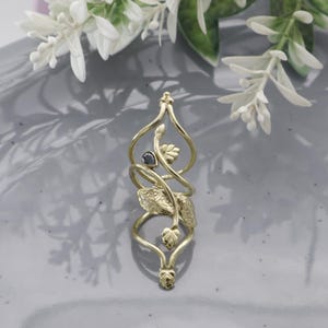 May include: A gold-colored pendant with an intricate, swirling design. The pendant features leaf and vine details, with a small, dark gemstone at the center. The overall shape is elongated and elegant, suggesting a nature-inspired theme.