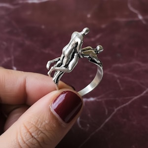 Puede incluir: Un anillo plateado con un diseño esculpido de dos figuras en un abrazo. El anillo tiene un acabado pulido y se sostiene sobre un fondo oscuro y estampado. El diseño del anillo es el punto focal.