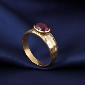 Peut inclure: Une bague dorée avec un anneau texturé martelé et une pierre précieuse rouge foncé ovale sertie en lunette. La bague est mise en valeur sur un fond bleu foncé, soulignant les détails.