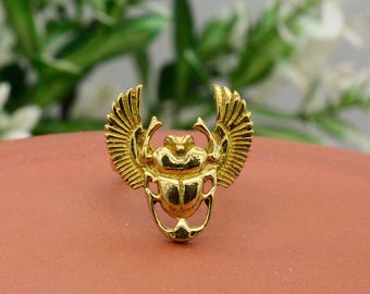 May include: A gold-coloured ring with a detailed scarab beetle design and outstretched wings. The ring is set against a reddish-brown surface, with a blurred green and white background.