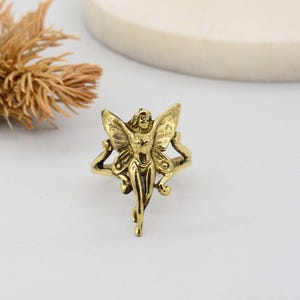 May include: A gold-colored ring featuring a detailed fairy design. The fairy has outstretched wings and is centered on the ring's band. The ring has an ornate, vintage aesthetic.