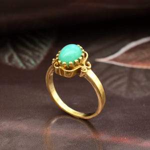 May include: A gold ring featuring a light green, oval-shaped gemstone. The gemstone is set in a decorative bezel setting. The ring's band is a simple, polished gold. The ring is a piece of jewelry.