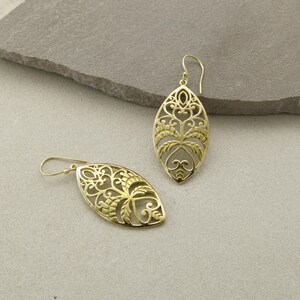 May include: A pair of gold-colored filigree earrings. Each earring has an elongated oval shape with intricate cut-out patterns. The design includes floral and leaf-like motifs, and a hook for pierced ears. The earrings are displayed on a neutral background.