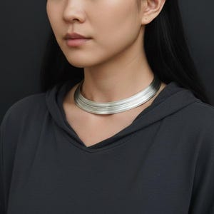 Puede incluir: Un collar gargantilla plateado con un diseño en capas. El collar se lleva sobre una sudadera con capucha gris oscuro. El fondo es de color gris oscuro liso.