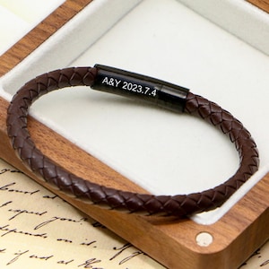 Peut inclure: Un bracelet en cuir tressé marron avec un fermoir noir. Le fermoir porte l'inscription "A&Y 2023.7.4". Le bracelet est présenté dans un écrin en bois avec un intérieur blanc.