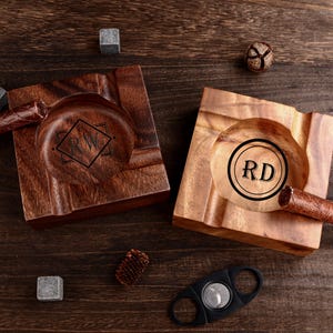 May include: Two wooden cigar ashtrays with personalized initials, one with "RW" in a diamond and the other with "RD" in a circle. A cigar cutter, cigar, and whiskey stones are also present.
