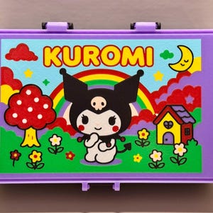 May include: A purple lunchbox featuring a colorful illustration of the character Kuromi. The design includes a rainbow, a mushroom, a house, and the word "KUROMI" in yellow letters. The lunchbox has a clasp closure.