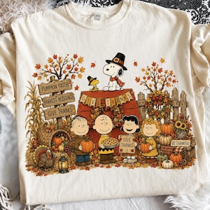 Puede incluir: Una camiseta color crema con un gráfico de Peanuts con temática de Acción de Gracias. El diseño muestra a Snoopy y Woodstock sobre una caseta de perro con hojas de otoño, calabazas, un pavo y los personajes Charlie Brown, Lucy y Sally.