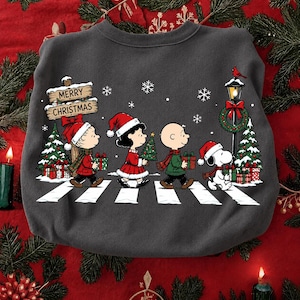 Puede incluir: Una sudadera gris oscuro de cuello redondo con un gráfico de los personajes de Peanuts cruzando un paso de cebra. Snoopy, Charlie Brown, Lucy y Linus llevan gorros de Papá Noel y regalos, junto a una farola y un cartel que dice MERRY CHRISTMAS.
