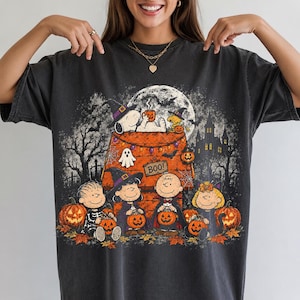 Puede incluir: Una camiseta de manga corta gris carbón con un estampado gráfico de los personajes de Peanuts con disfraces de Halloween. El diseño incluye a Snoopy sobre una caseta, calabazas, una casa encantada, murciélagos y el texto BOO!.