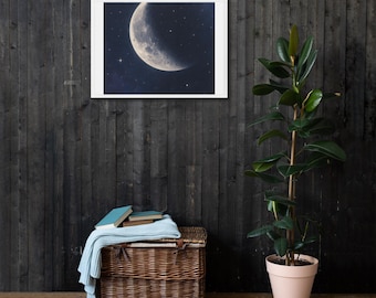 May include: A framed canvas print featuring a crescent moon and stars against a dark blue background. The artwork is displayed on a dark wood-paneled wall. A woven basket with a blue blanket and books sits below, with a potted plant to the right.