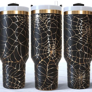 Könnte beinhalten: Drei schwarze Reisebecher mit einem goldenen Spinnennetz-Design. Jeder Becher hat einen schwarzen Griff, einen weißen Deckel und ein goldenes Band am oberen Rand. Die Becher sind für heiße oder kalte Getränke konzipiert.
