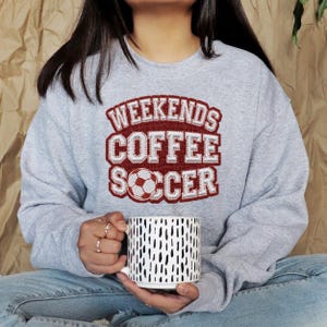 May include: A light gray sweatshirt with the words "WEEKENDS COFFEE SOCCER" in a red and white design. A white mug with black dashes is held in the hands.