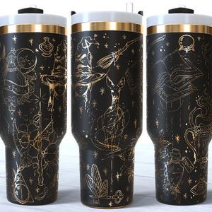 May include: A black stainless steel tumbler with a handle, featuring a gold-toned line art pattern of magical items including a witch hat, broomstick, potion bottles, a crystal ball, an open book, a wand, and stars on a matte black background.