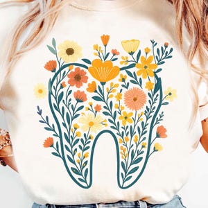 Peut inclure: Sweat-shirt crème avec un motif de dent floral. La dent est soulignée en bleu sarcelle et remplie de fleurs jaunes, oranges et pêche et de feuilles vertes. Le motif est centré sur le devant du t-shirt.