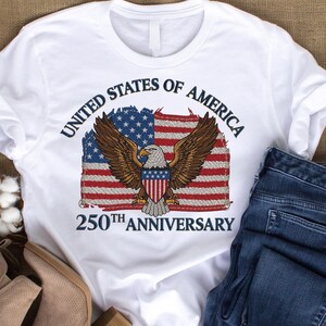 以下が含まれることがあります： 愛国的なデザインの白いTシャツ。グラフィックには、ワシ、アメリカ国旗、テキスト「UNITED STATES OF AMERICA」と「250TH ANNIVERSARY」が含まれています。このTシャツは、青いジーンズと組み合わせています。