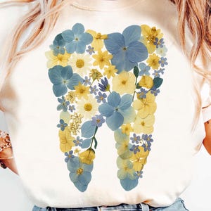 Peut inclure: T-shirt crème avec un motif de dent composé de fleurs bleues et jaunes. L'arrangement floral comprend des hortensias, des marguerites et d'autres petites fleurs. Le t-shirt a un col rond et des manches courtes.