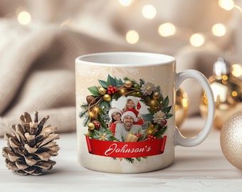 Könnte beinhalten: Eine weiße Keramiktasse mit einem Familienfoto in einem Weihnachtskranz-Design. Der Kranz enthält Tannennadeln, Beeren und goldene Ornamente. Ein rotes Banner am unteren Rand trägt die Aufschrift "Johnson's". Die Tasse steht vor einem verschwommenen Hintergrund aus Lichtern.