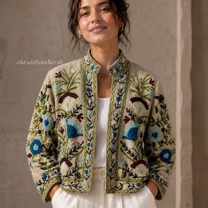 May include: A beige jacket featuring intricate floral embroidery in shades of blue, green, and dark red. The jacket has a mandarin collar, long sleeves, and an open front design. The text zikraanhandicraft is visible on the upper left side.
