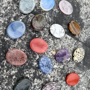 Pode incluir: Uma coleção de pedras polidas em forma oval, em várias cores e padrões. As pedras incluem tons de azul, rosa, vermelho, verde e castanho, com texturas e marcas únicas. As pedras estão dispostas sobre uma superfície cinza texturizada.