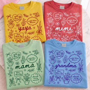 Könnte beinhalten: Vier farbige T-Shirts mit floralen Designs und Text. Die Shirts sind gelb, rot, grün und blau. Jedes Shirt hat ein anderes Wort: "yaya", "mimi", "mama" und "Oma". Die Shirts haben einen Rundhalsausschnitt und kurze Ärmel.
