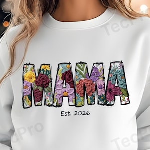 Könnte beinhalten: Weißes Sweatshirt mit dem Wort "MAMA" in großen, floral gefüllten Buchstaben. Die Buchstaben sind schwarz umrandet und mit verschiedenen bunten Blumen und Grünzeug gefüllt. Unter dem Wort "MAMA" steht der Text "Est. 2026".