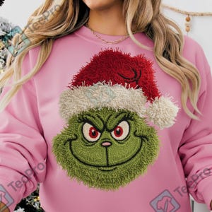 May include: Pink sweatshirt featuring a green, textured Grinch face wearing a red Santa hat with white trim. The Grinch has a mischievous grin and red eyes. A festive holiday-themed apparel item.