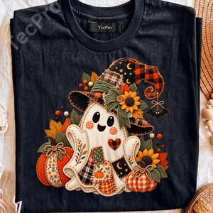 May include: A black t-shirt with a crew neckline and a TecPro label, featuring a graphic of a white ghost wearing a witch hat decorated with sunflowers, plaid patterns, and stars, flanked by orange pumpkins and autumn leaves on a dark background.