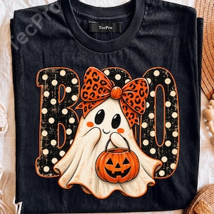 May include: A black short-sleeve t-shirt featuring a graphic of a white ghost wearing an orange and black leopard print bow, holding an orange jack-o'-lantern. The word BOO is printed in large black letters with white polka dots behind the ghost.